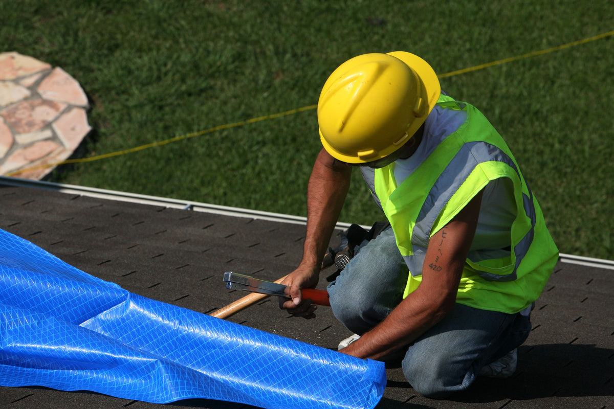 Roofer installing a temporary blue tarp repair on a residential roof after storm damage